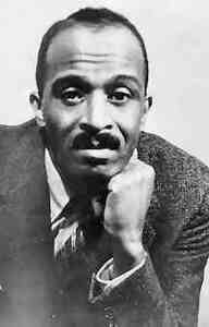 J. Saunders Redding, a Black man with a mustache, rests his chin on his left fist and looks into the camera. He is wearing a suit, collared shirt, and striped tie.