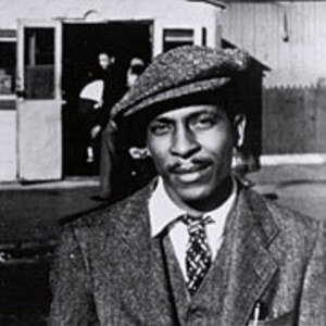 Andy Razaf, a Black man with a mustache, stands in front of a building wearing a tweed flat cap with a matching coat