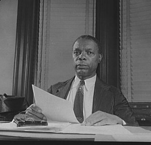 William Pickens, a Black man, sits at a desk holding a piece of paper and looking into camera.
