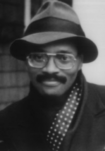 Larry P. Neal, a Black man with a mustache, wears a fedora, aviator glasses, jacket, and polka dot scarf smiles softly.