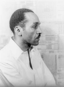 A Black man, Willard Motley's, profile. He wears a white shirt and stands in front of a wall covered in geometric patterns.